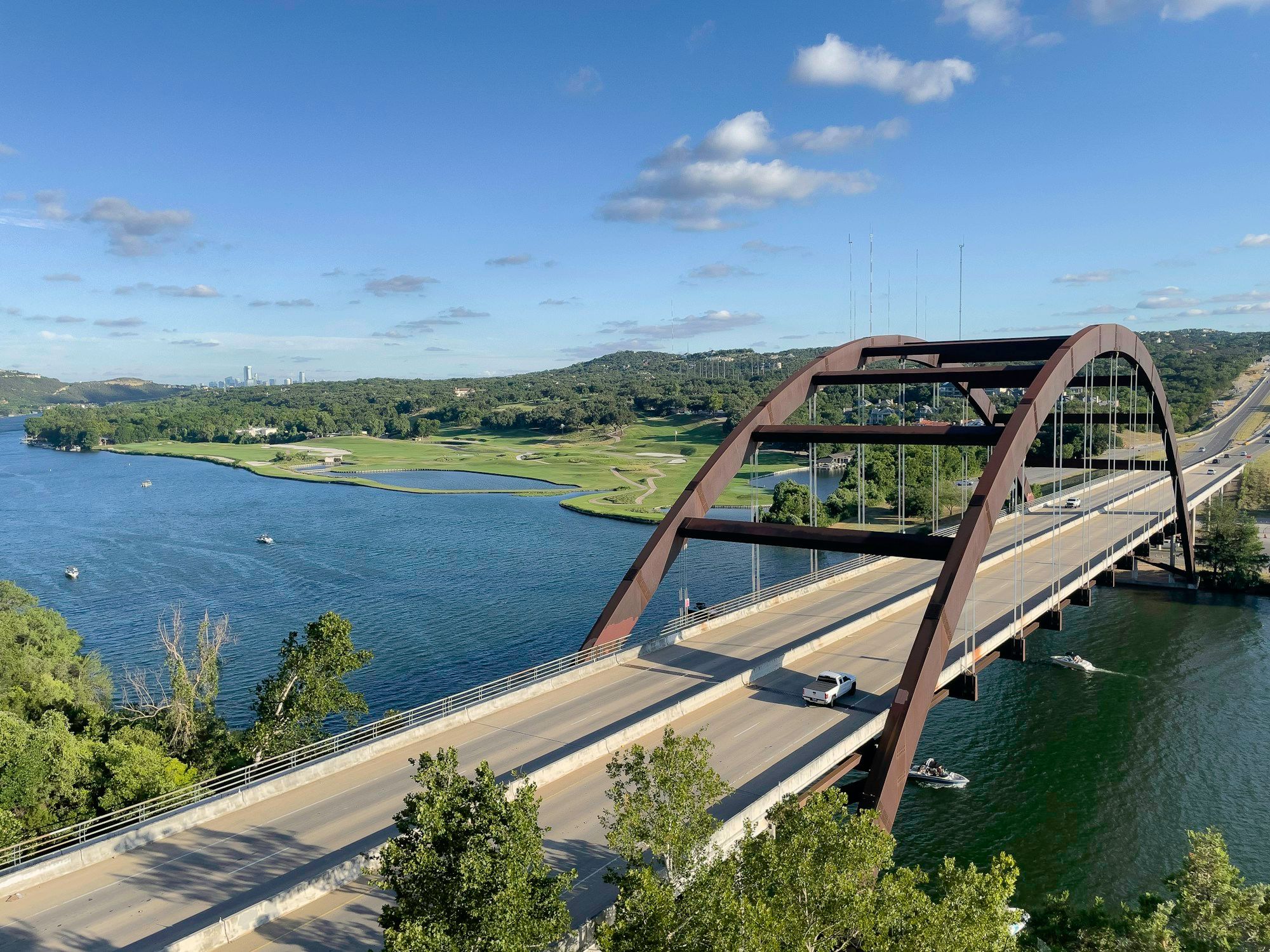 The rust-colored arch of the Pennybacker Bridge over Lake Austin in west Austin, with green hills and a golf course beyond