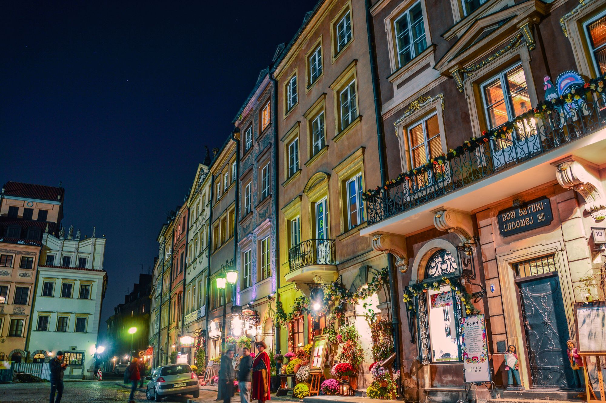 A lit street of colorful townhouses with flower stalls in Warsaw's Old Town in the evening.