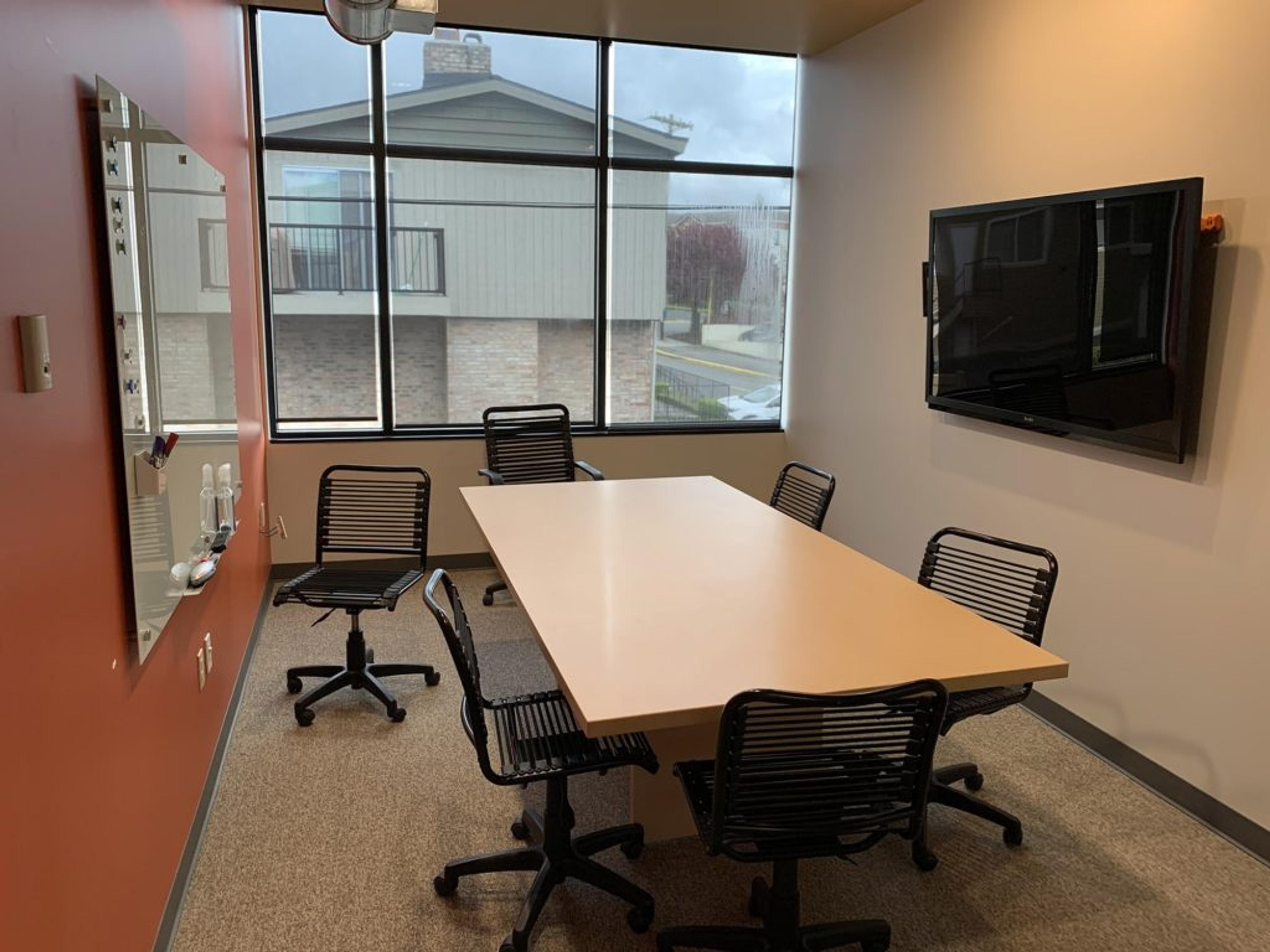 An empty conference room with table and chairs around it with a tv on one wall and a white board on the opposite side. Big windows with shades pulled down.