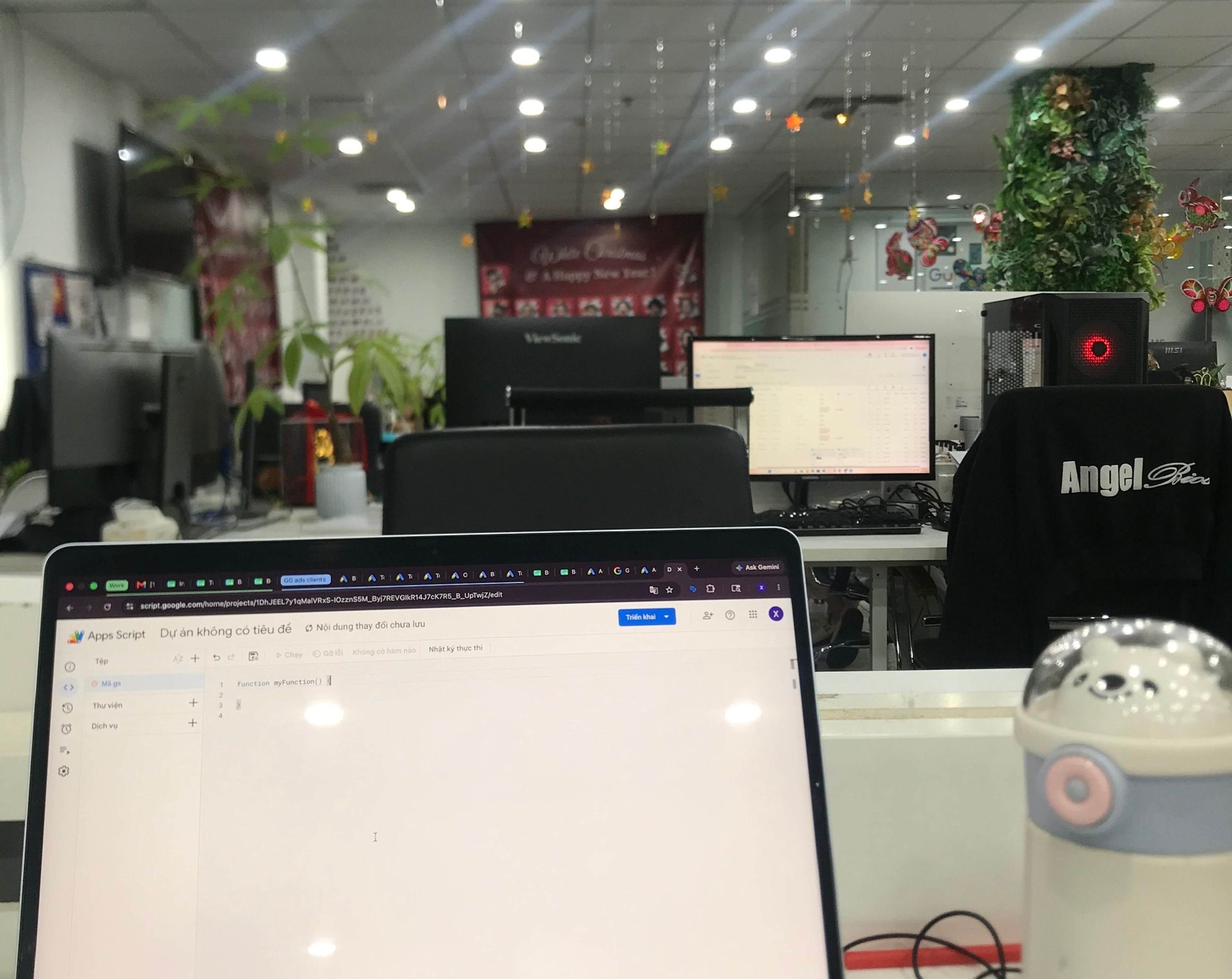 An open laptop on a desk in a brightly lit office, a Google Apps Script editor on screen