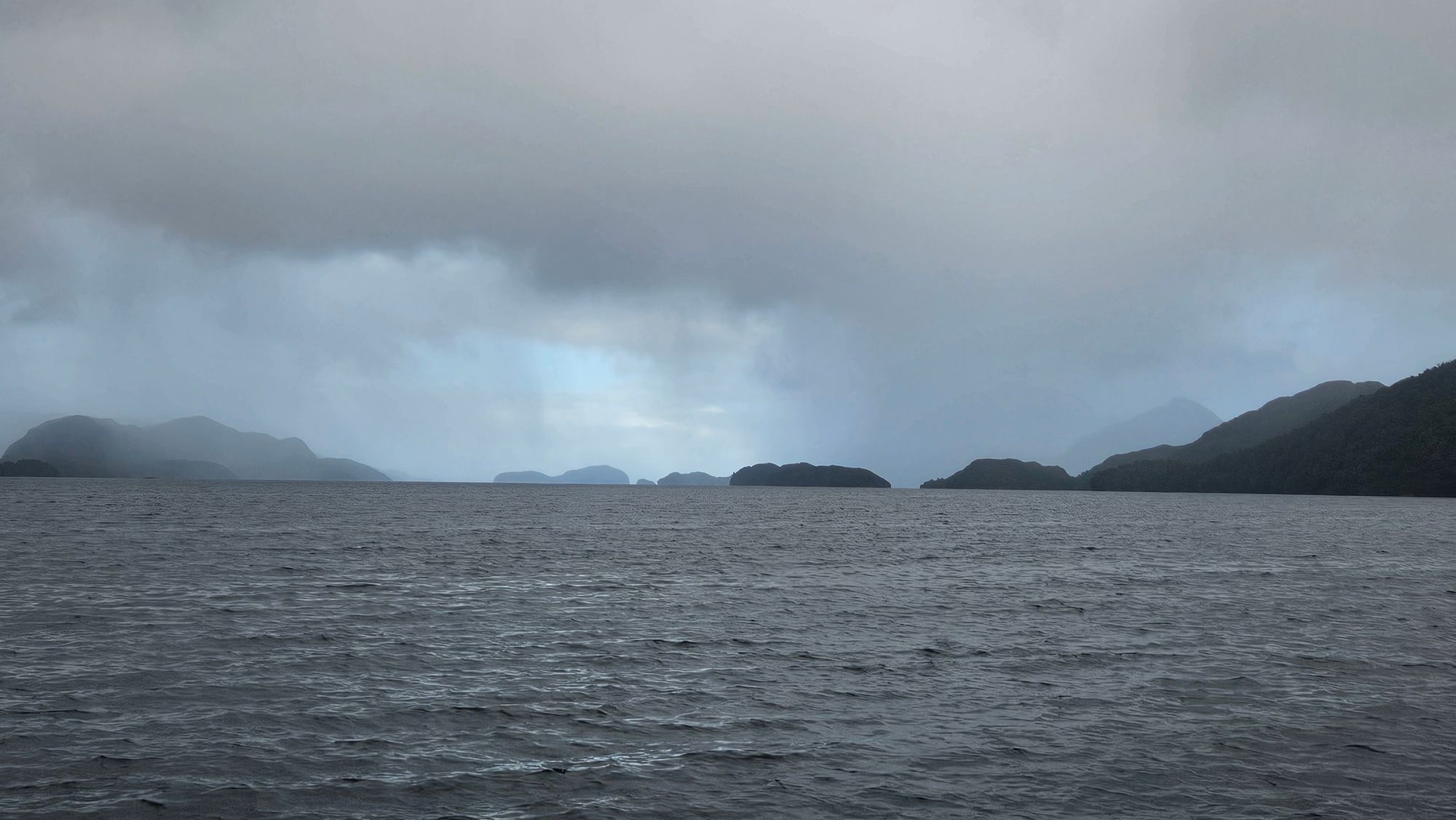 Dusky Sound approach in heavy overcast — island silhouettes emerging from rain and mist across open water