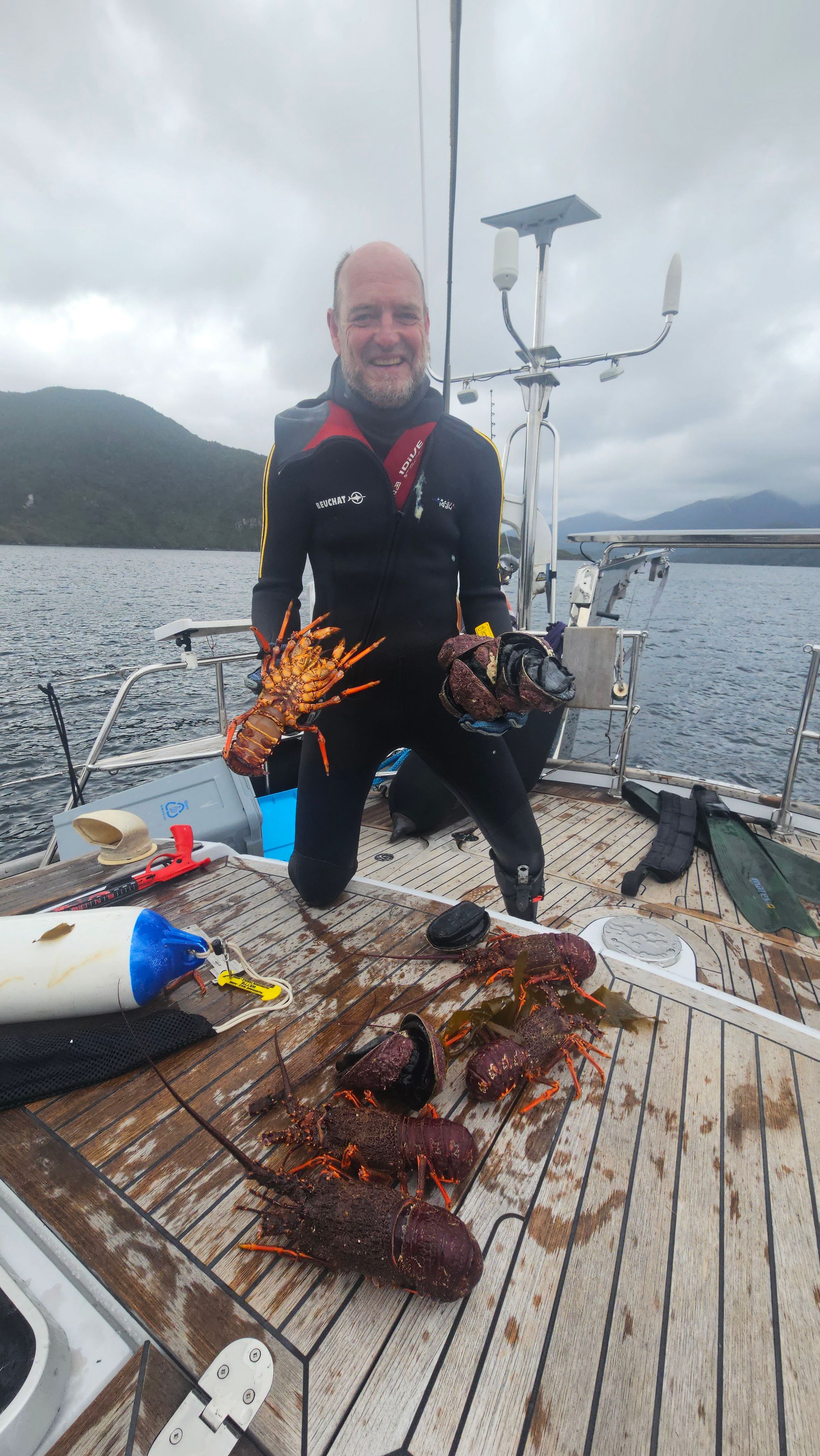 Greg in wetsuit on Matariki III's deck holding up a large crayfish with paua, more crayfish laid out in front