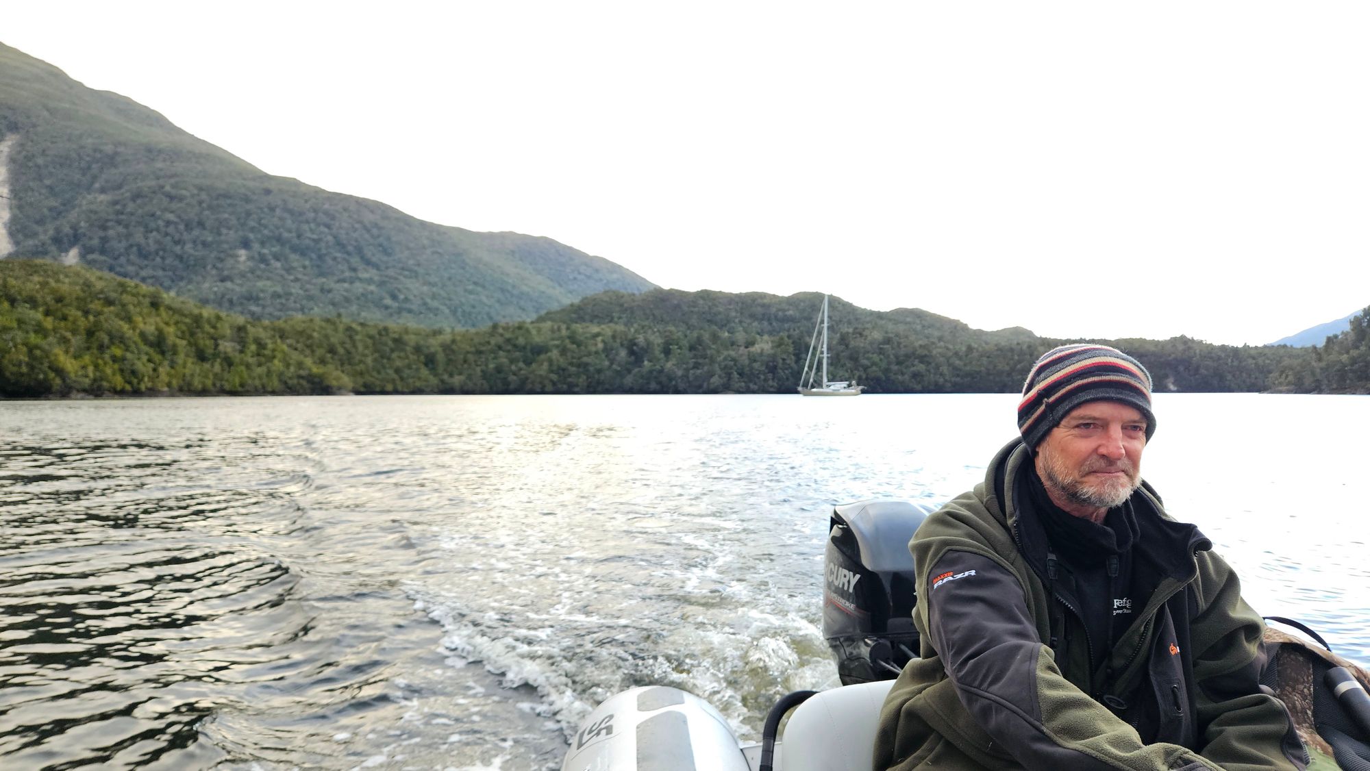 Bruce at the helm of the tender on Lake Cove, Matariki III visible at anchor in the background against the forested mountains