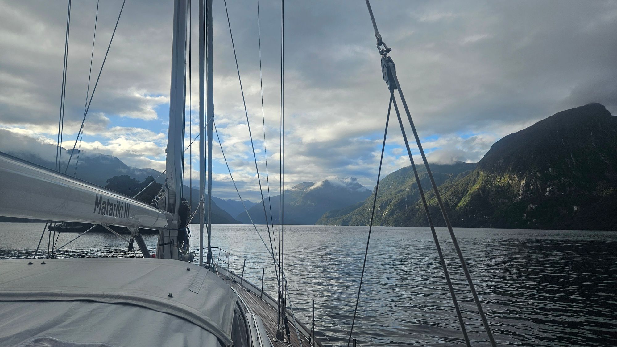 Matariki III at anchor with washing drying on the lifelines, Dusky Sound's forested cliffs and rock faces behind