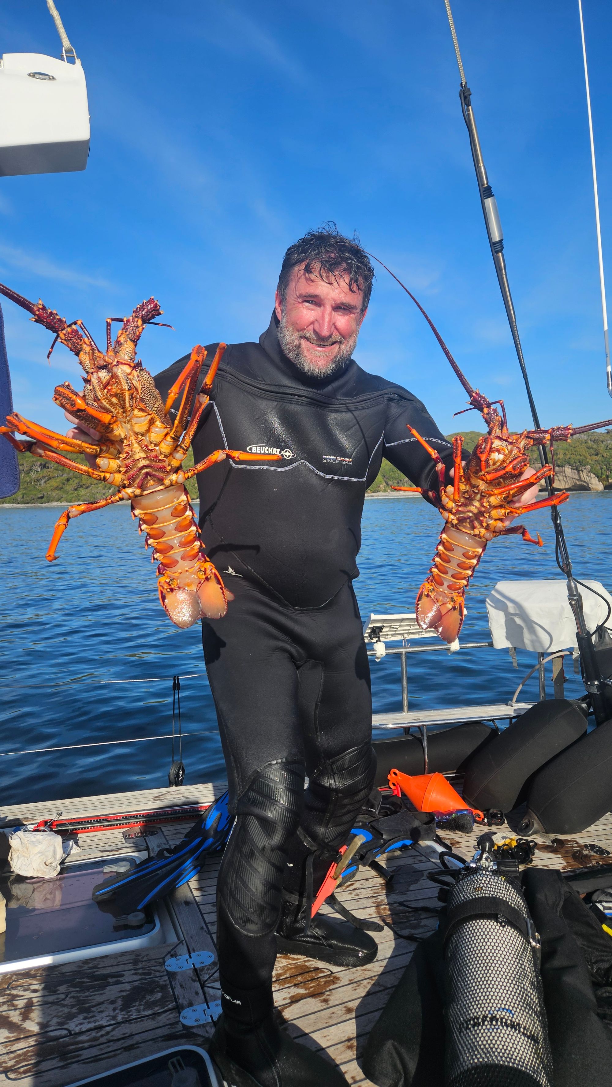 Greg in wetsuit on Matariki III's deck holding two enormous southern rock lobsters, one in each hand, after a dive in Chalky Inlet