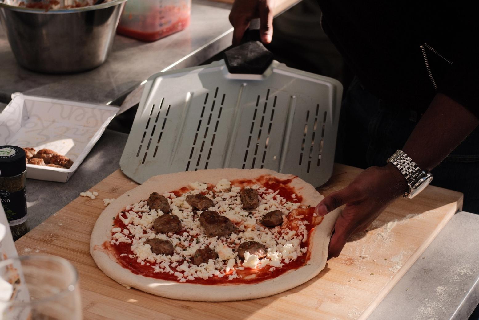 En person gjør klar en ubakt pizza med tomatsaus, ost og kjøttboller på en trefjøl, mens en pizzaspade holdes klar.