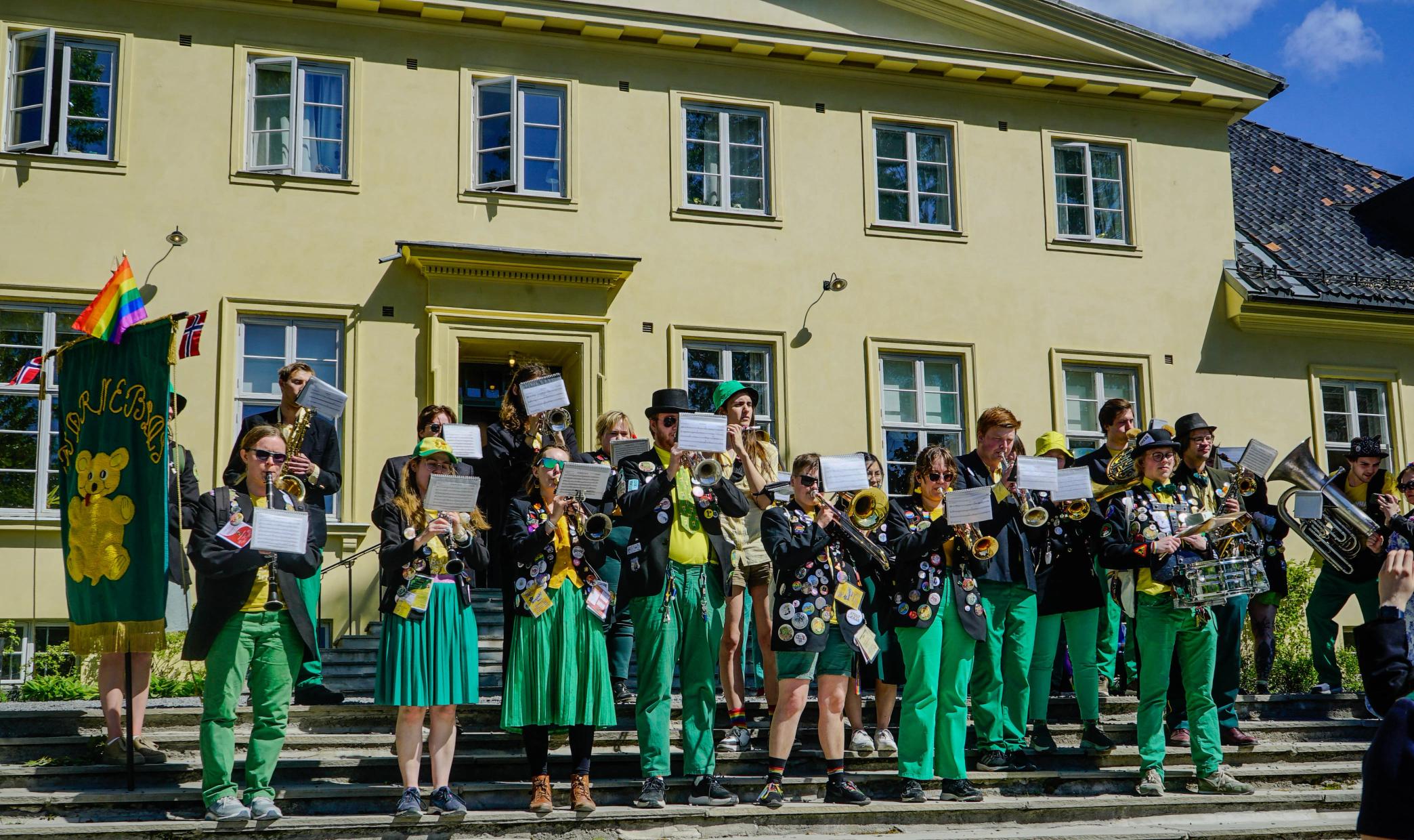 Et studentorkester i grønne og svarte klær spiller instrumenter på trappen foran en gul bygning, mens de holder noter og et grønt banner med en bamse. Norske flagg og et regnbueflagg vaier ved siden av dem i solskinnet.