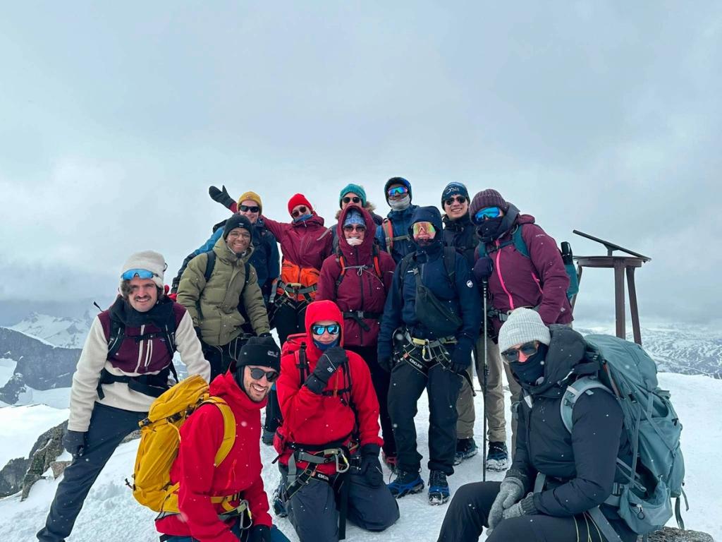 A group of mountaineers in colorful winter gear pose together on a snowy summit beneath an overcast sky.