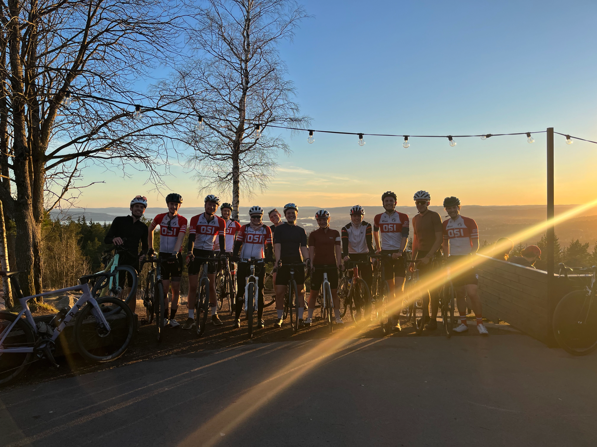A group of cyclists in matching jerseys stand with their bikes on a hilltop at sunset, with bare trees, string lights, and a scenic valley in the background.