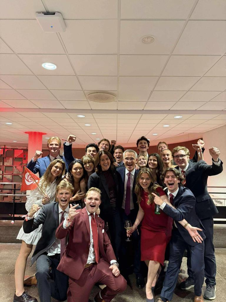 En stor gruppe unge voksne i pene klær jubler og smiler mot kameraet innendørs, flere med røde antrekk og et lite rødt flagg. Stemningen er festlig og feirende.