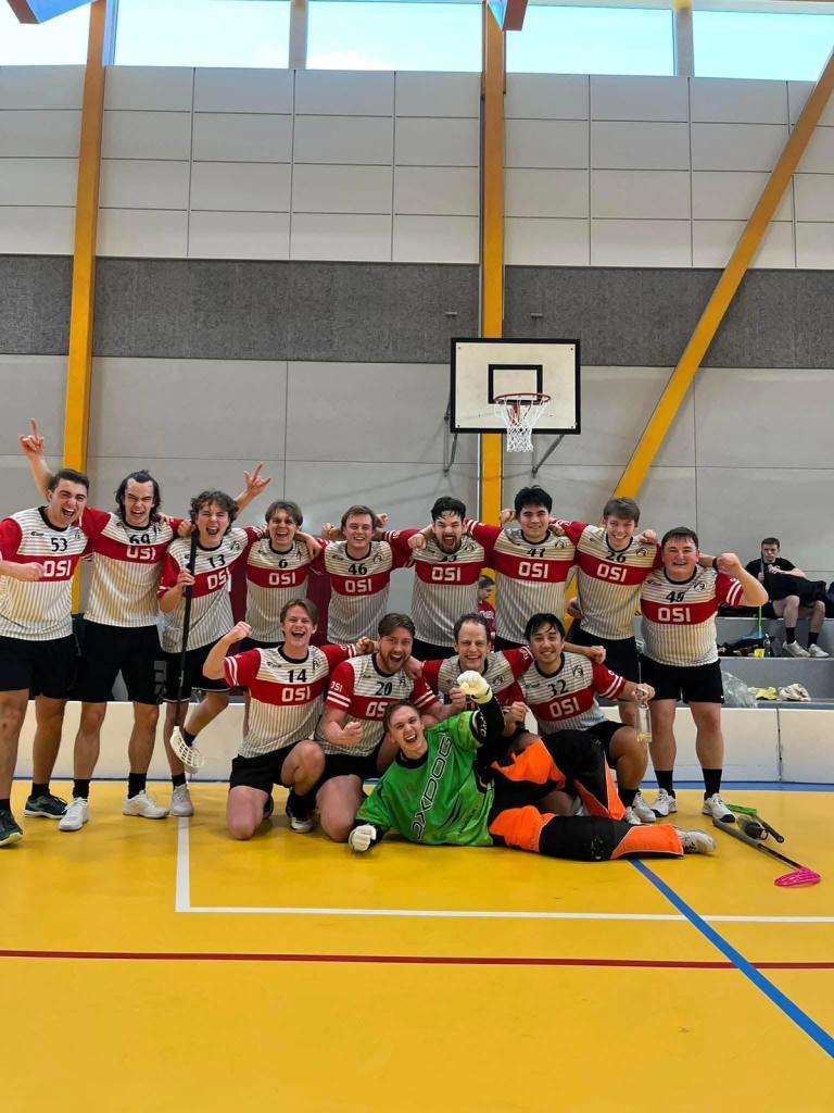 Et innebandylag i røde og hvite drakter jubler og poserer sammen på en gul innendørsbane foran en basketballkurv, med målvakten liggende foran i grønt og oransje utstyr.