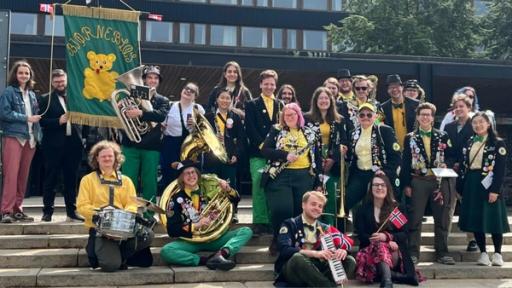 Et studentorkester i gule, grønne og svarte antrekk poserer på en trapp med instrumenter, norske flagg og en grønn fane med en gul bamse.
