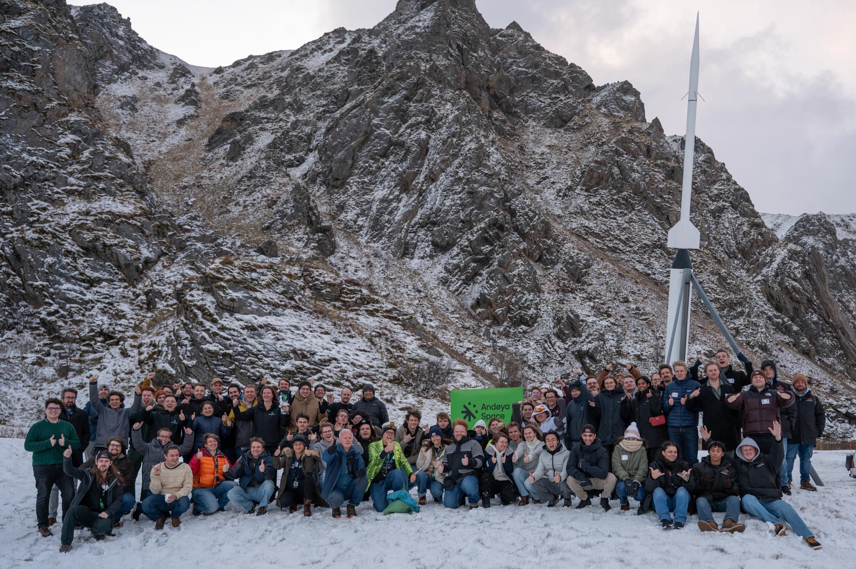 En stor gruppe mennesker poserer i snøen foran en rakett og bratte, snødekte fjell, med et skilt fra Andøya Space i midten.