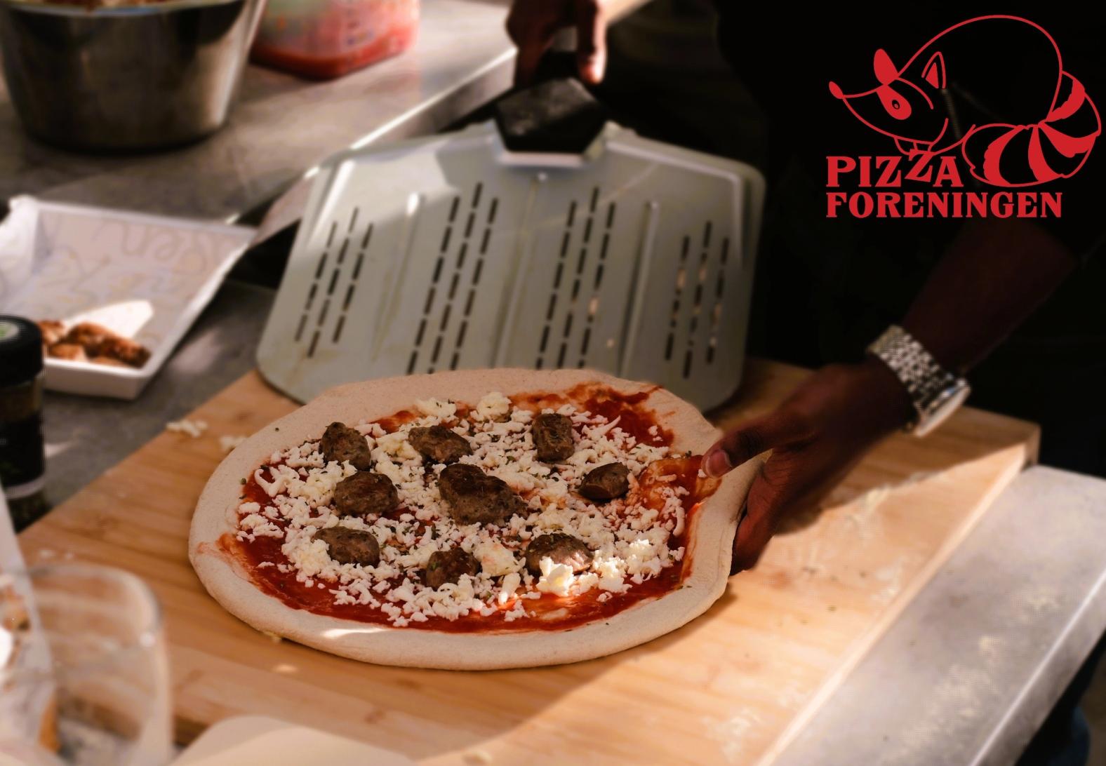 En person gjør klar en pizza med tomatsaus, ost og kjøttboller på en trefjøl. Pizza Foreningen-logoen vises øverst til høyre.