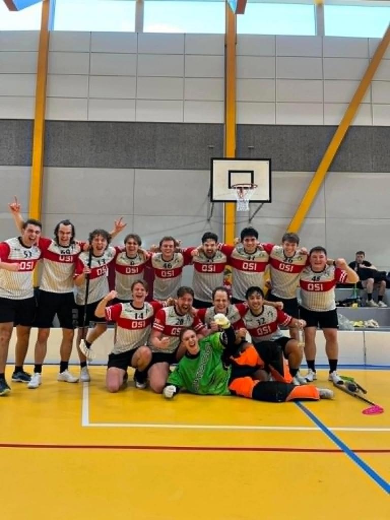 Et innebandylag i røde og hvite drakter jubler og poserer sammen på en gul hallflate foran en basketballkurv, med målvakten i grønn og oransje utstyr liggende foran laget.