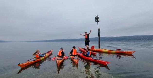En gruppe kajakkpadlere med oransje redningsvester samles på rolig vann, mens én person står oppreist i en kajakk med armene utstrakt.