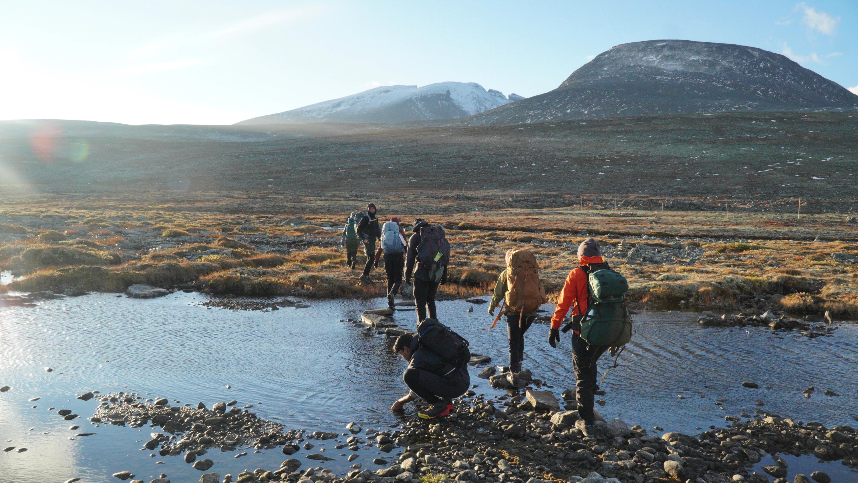 En gruppe fotturister med store ryggsekker krysser en grunn bekk over steiner i et åpent fjellandskap med snødekte topper i bakgrunnen.