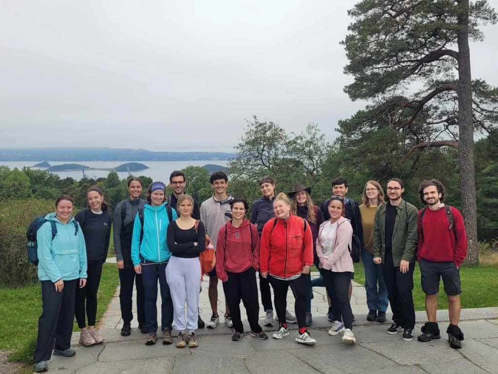En gruppe mennesker poserer sammen på et utsiktspunkt omgitt av trær, med fjorden og øyer i bakgrunnen under en overskyet himmel.