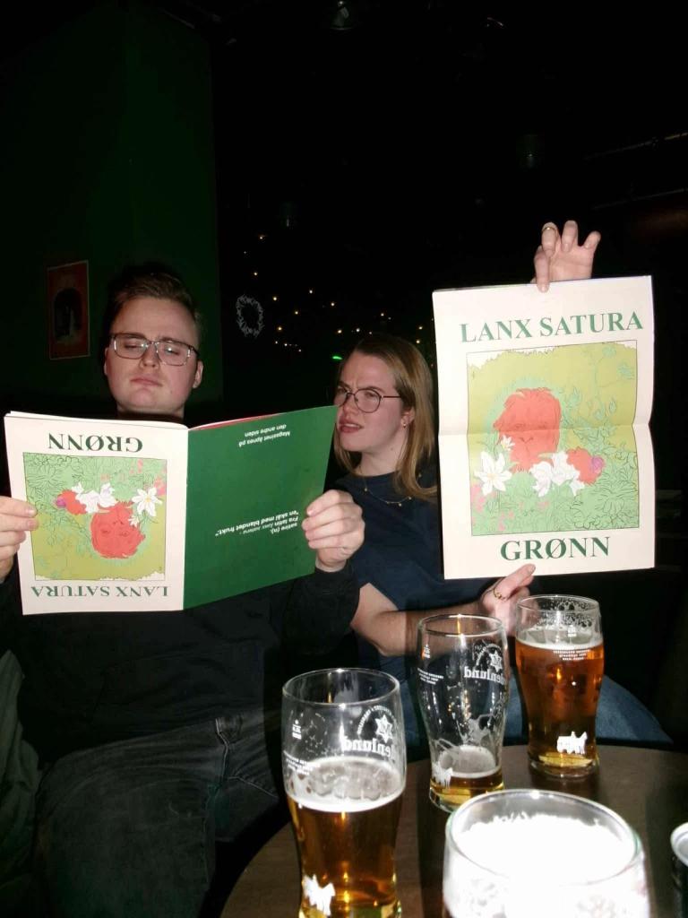 To personer sitter ved et bord med ølglass i en mørk bar og holder opp store, illustrerte menyer med teksten «Lanx Satura Grønn».