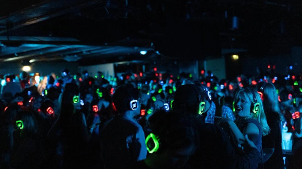 En stor folkemengde danser på en mørk klubb med fargerike, lysende hodetelefoner på hodet under en stille-disco.