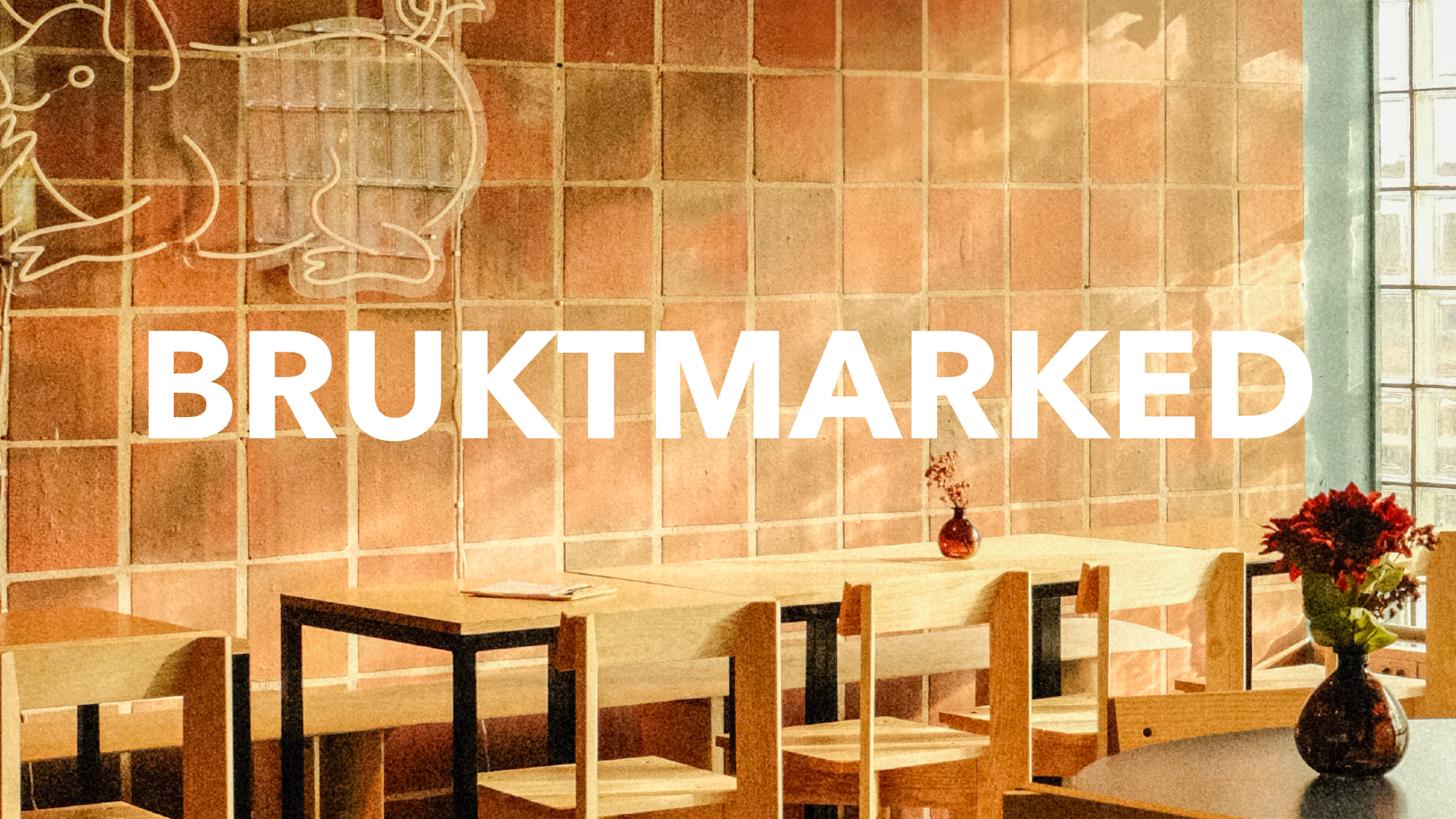 Lyst, retroinspirert kafélokale med enkle trebord og stoler, blomster i vaser på bordene og stor hvit tekst «BRUKTMARKED» over et rutete murveggmønster.