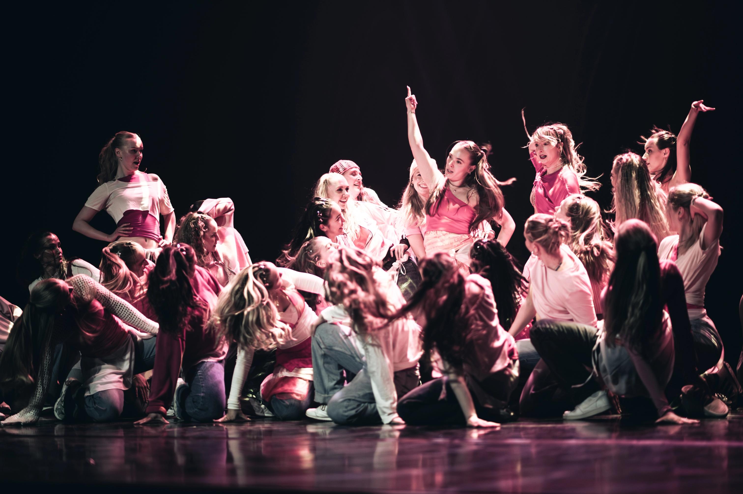 A large group of dancers in pink and white outfits perform an energetic routine on stage, with one dancer in the center pointing upward as others pose dynamically around her.