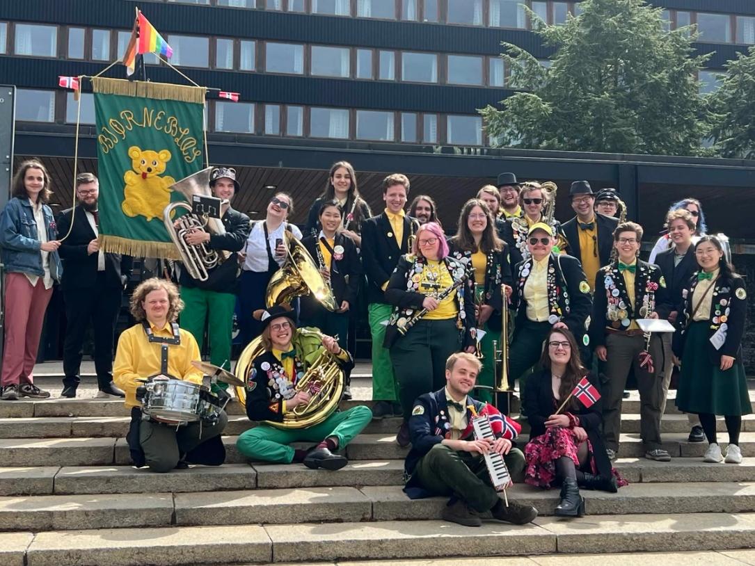 En stor gruppe glade musikanter i fargerike uniformer og med instrumenter poserer på en trapp foran en bygning, med et grønt «Bjørneblæs»-banner og flere norske flagg synlige.