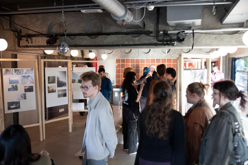Besøkende ser på fotografier og snakker sammen under en utstilling i et moderne lokale med synlige ventilasjonsrør og en diskokule i taket.