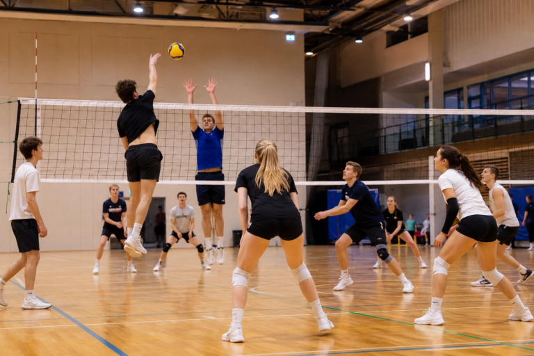 En gruppe unge voksne spiller volleyball innendørs, der en spiller hopper høyt for å smashe ballen over nettet mens motstanderen forsøker å blokkere.