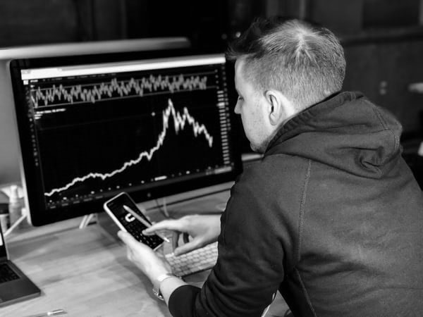 Black-and-white photo of a person viewing a stock chart on a monitor while holding a phone.