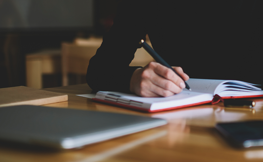 Close-up of a hand writing in a notebook on a wooden desk.