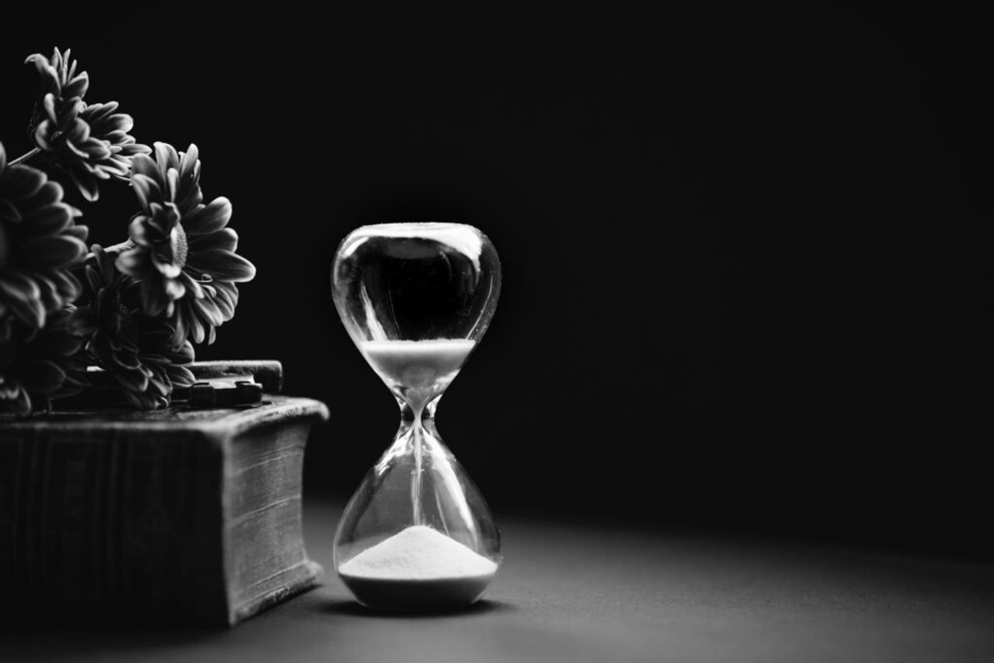 Black-and-white still life of an hourglass beside a book and flowers against a dark background.