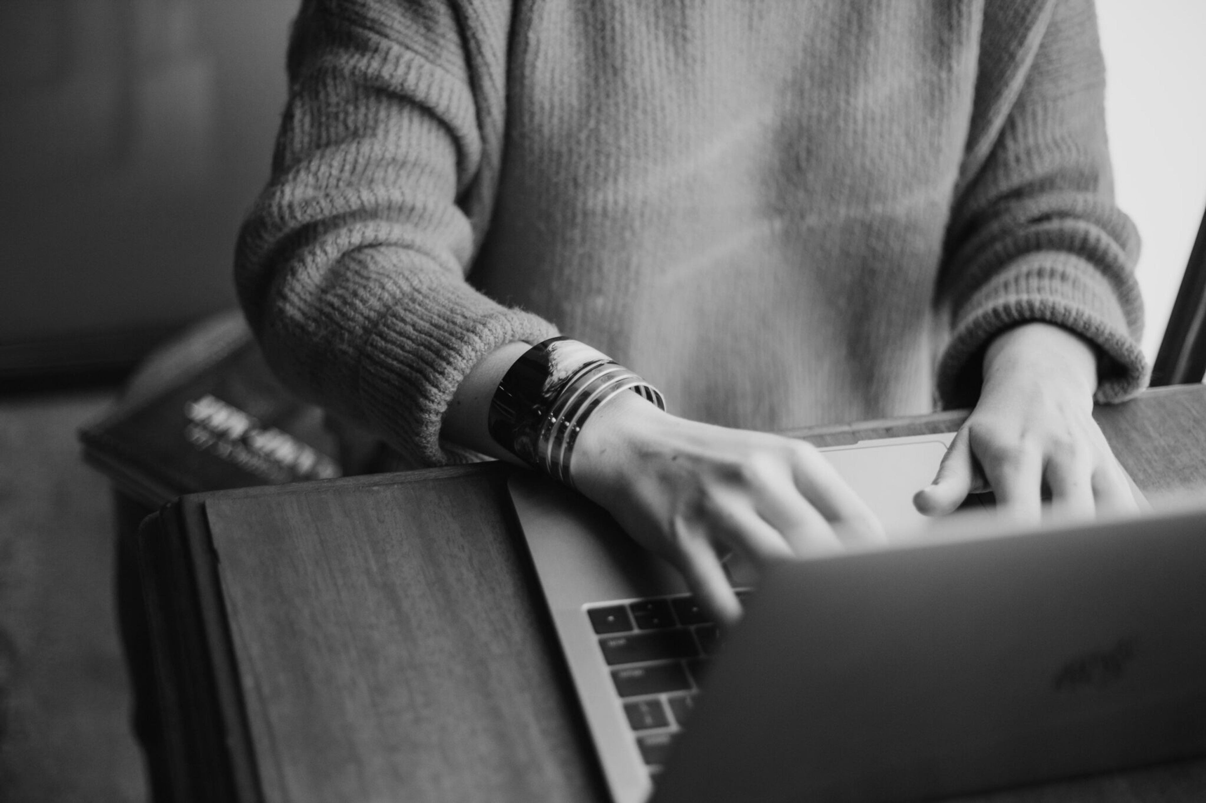 Black-and-white close-up of a person typing on a laptop at a wooden desk.