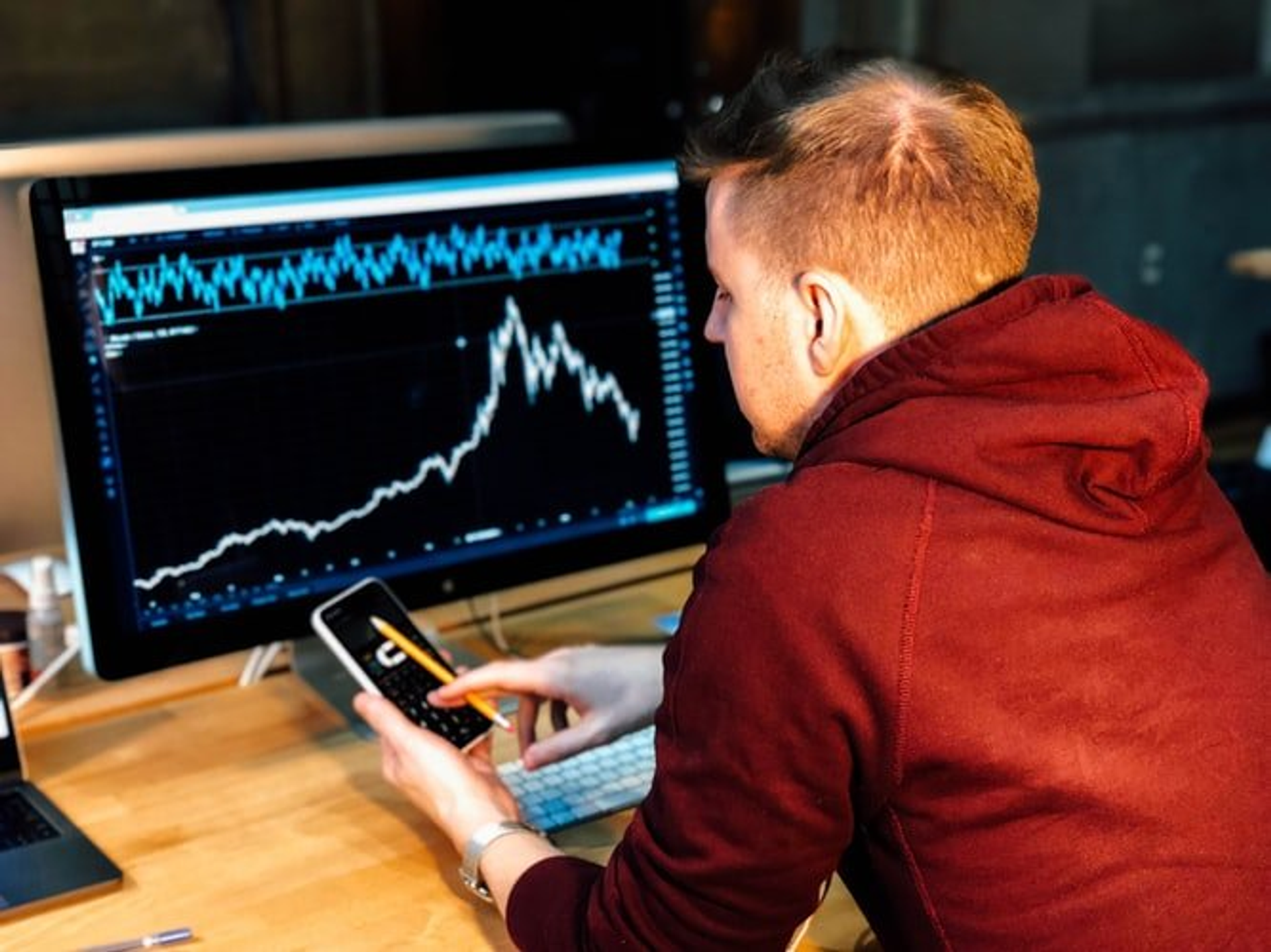 Trader reviewing a price chart on a desktop monitor while holding a smartphone.