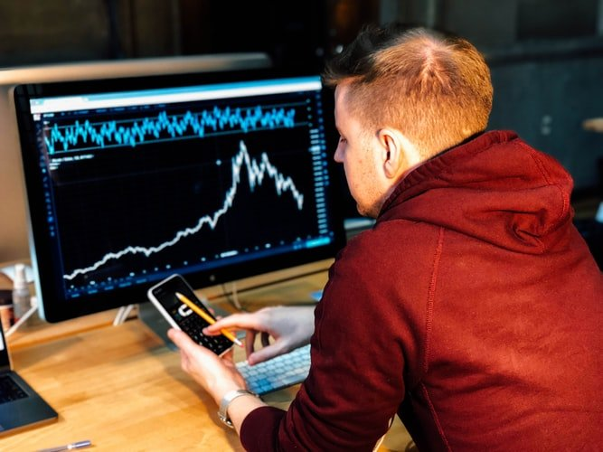 Trader reviewing a price chart on a desktop monitor while holding a smartphone.