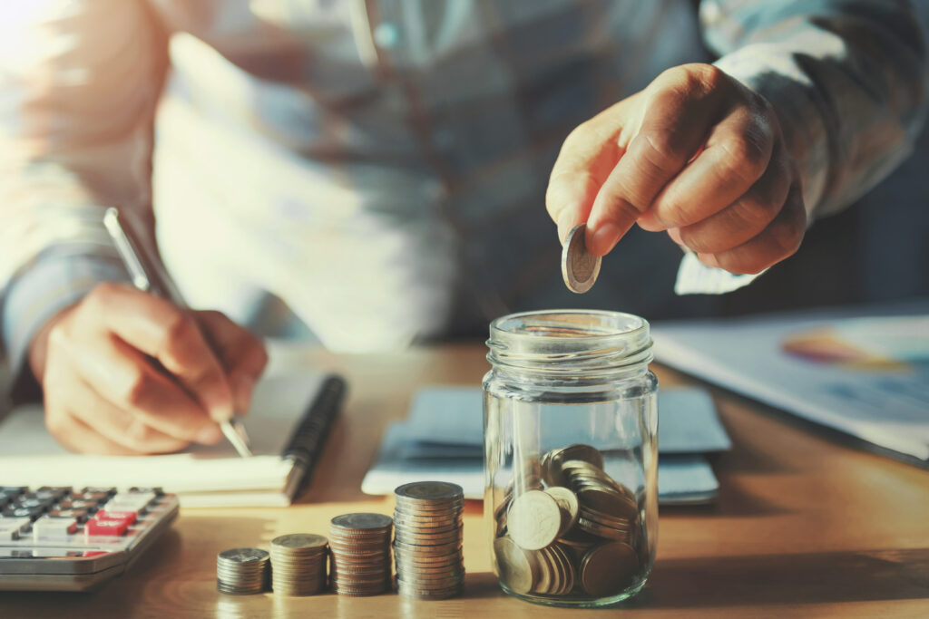 Person placing coins into a glass jar on a desk with a calculator and paperwork.
