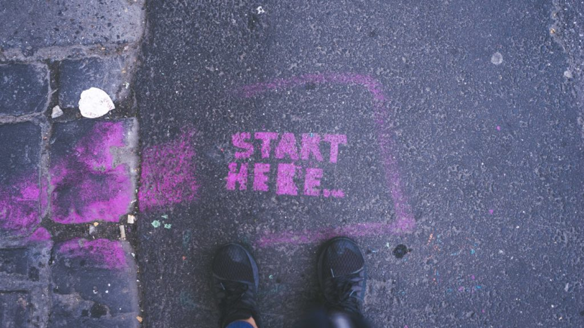 Chalk text reading “Start Here” on pavement with shoes visible.