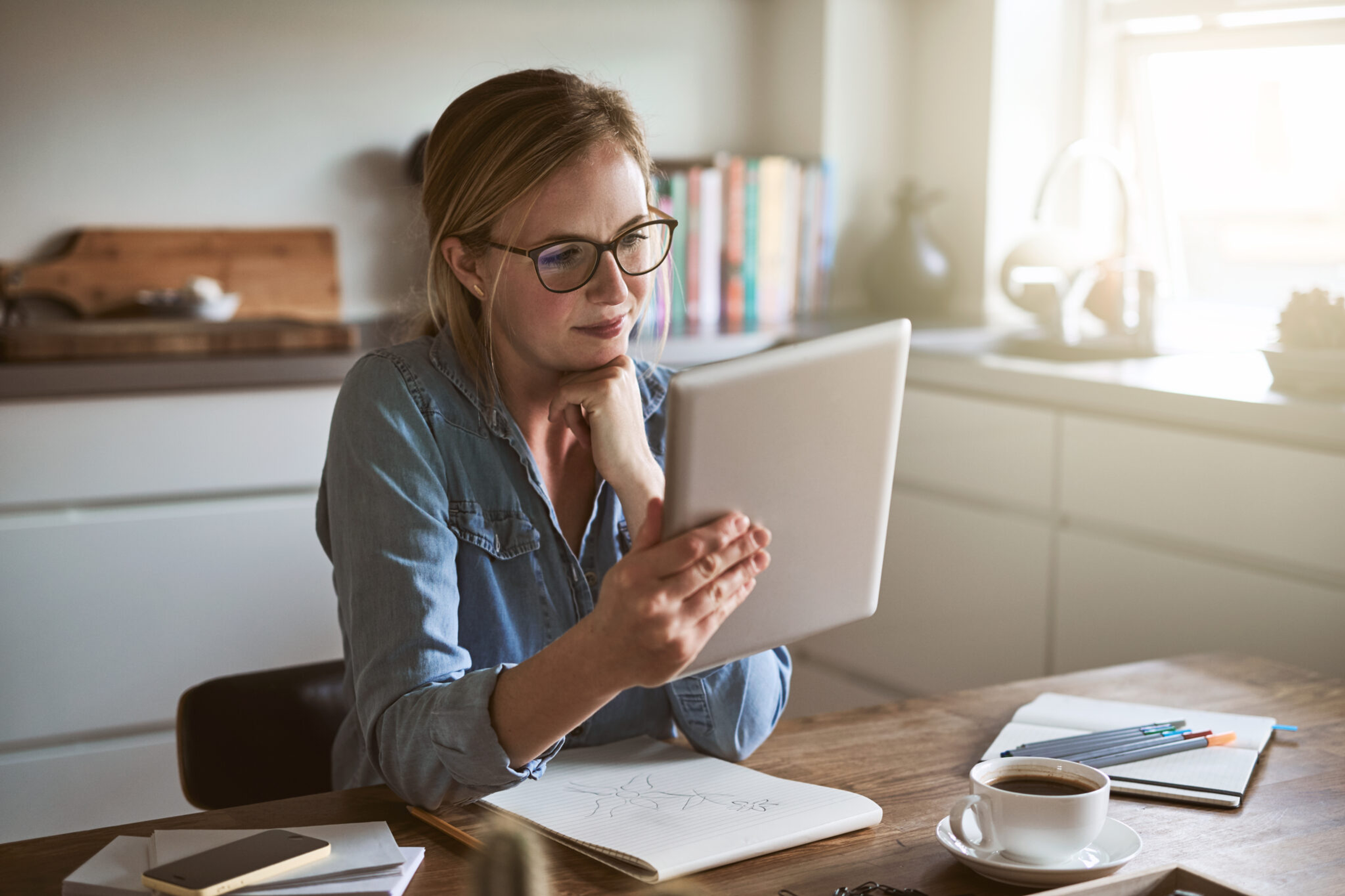 Woman with glasses using tablet at desk with notebook and coffee