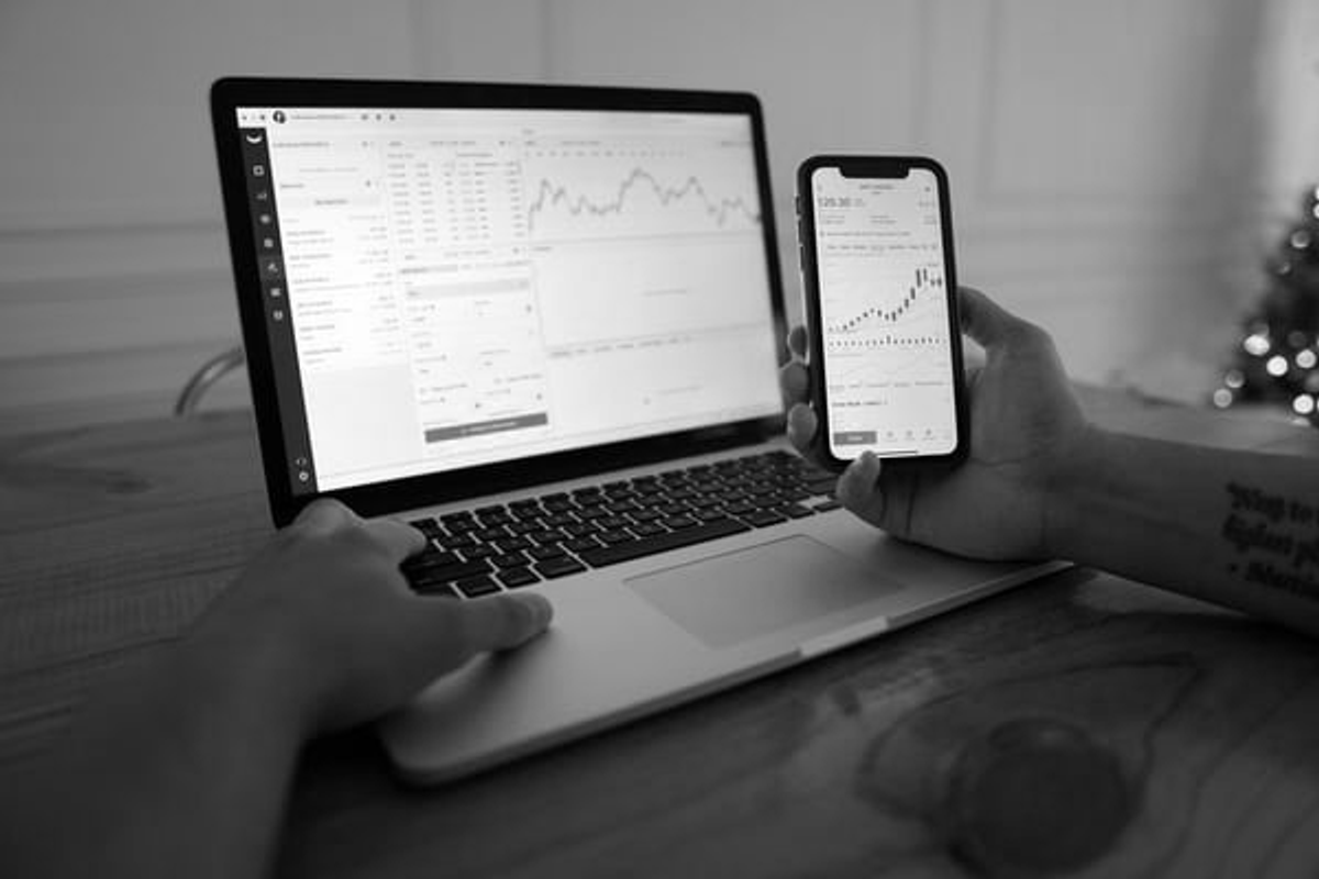 Person viewing trading charts on a laptop while holding a smartphone with a market graph.