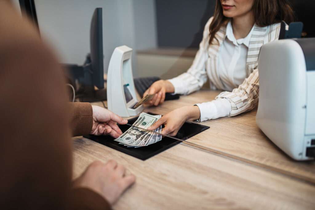 Bank teller handing cash to a customer through a glass partition.