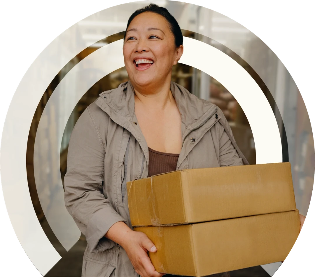 Young Asian woman wearing a tan jacket over a brown shirt carrying two small cardboard boxes looking off to the side of the camera and smiling.