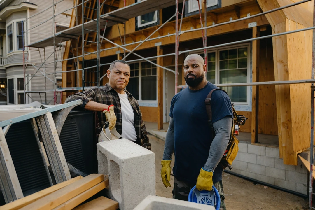 Two Black male construction workers with serious expressions standing in front of a house undergoing construction facing the camera.