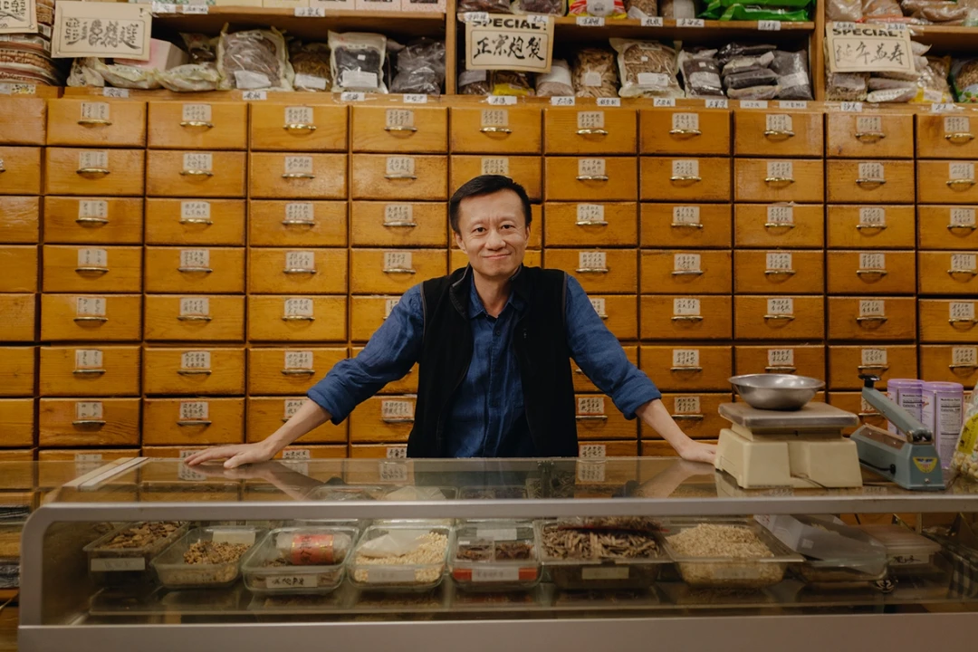 Middle-aged Asian man standing behind a glass counter inside a shop selling herbs and teas.