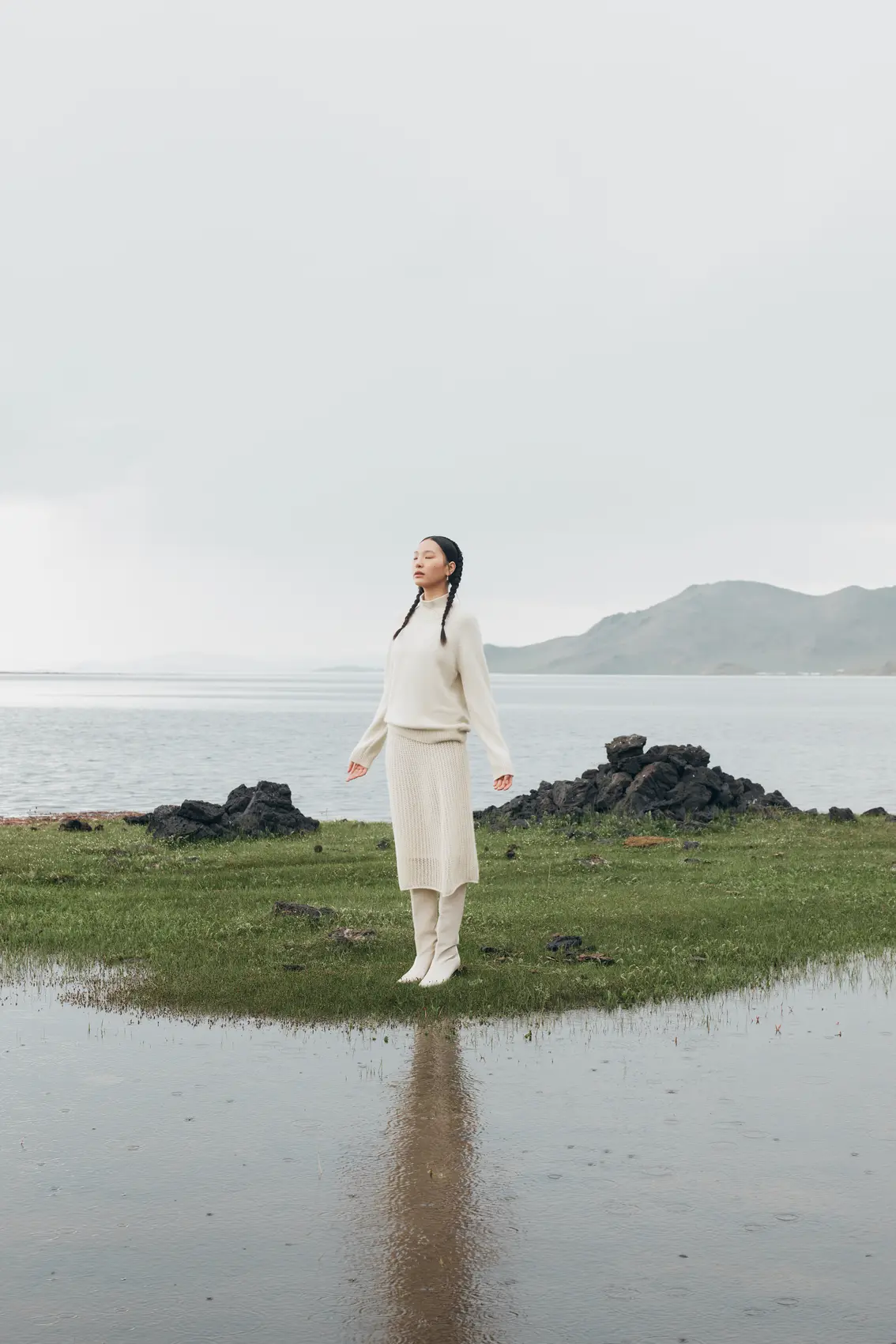 Front view of a woman standing on a little grass land in the middle of a lake wearing white cashmere sweater and skirt | GOBI Cashmere