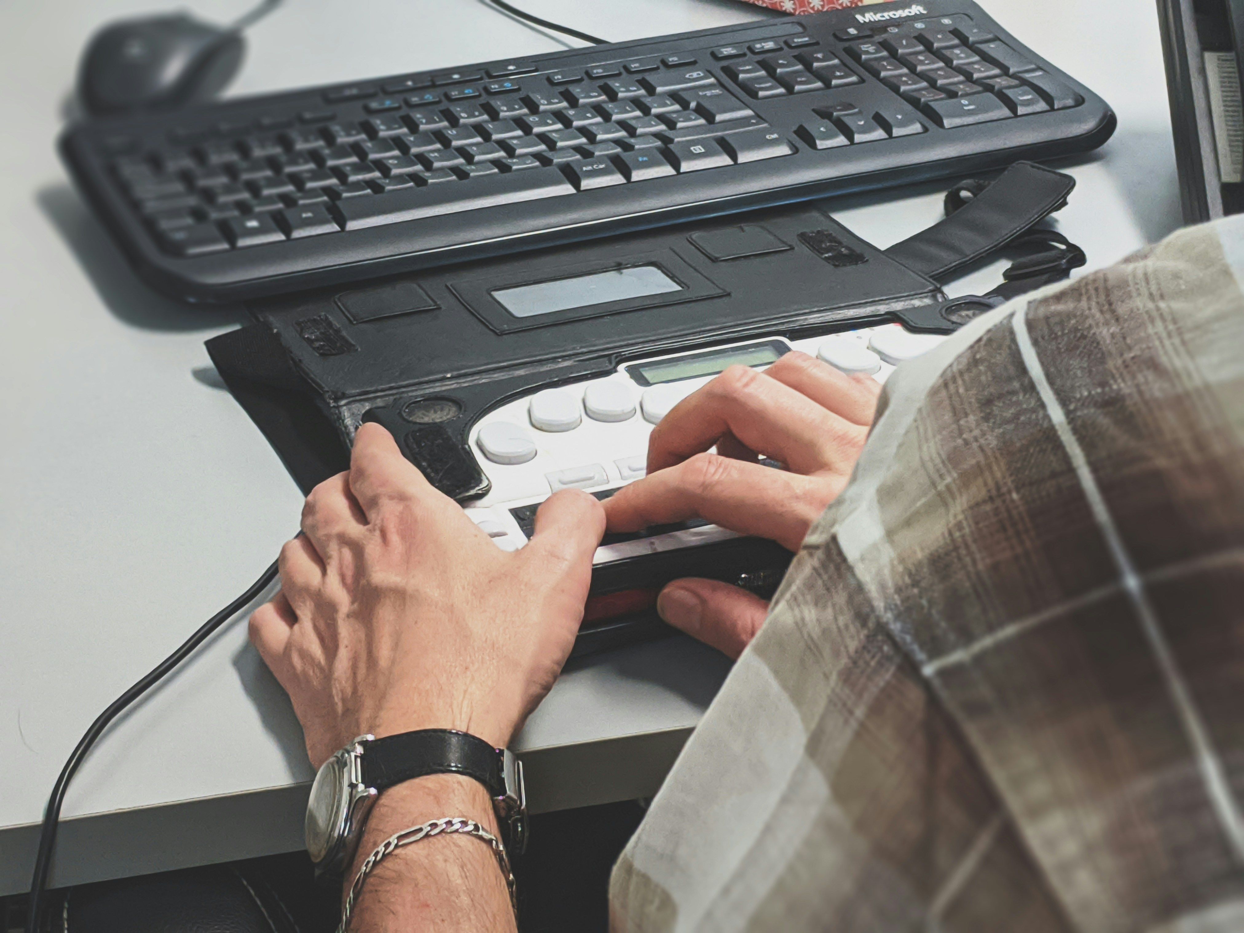 A person using a refreshable braille display connected to a desktop computer, with a standard keyboard visible in the background.