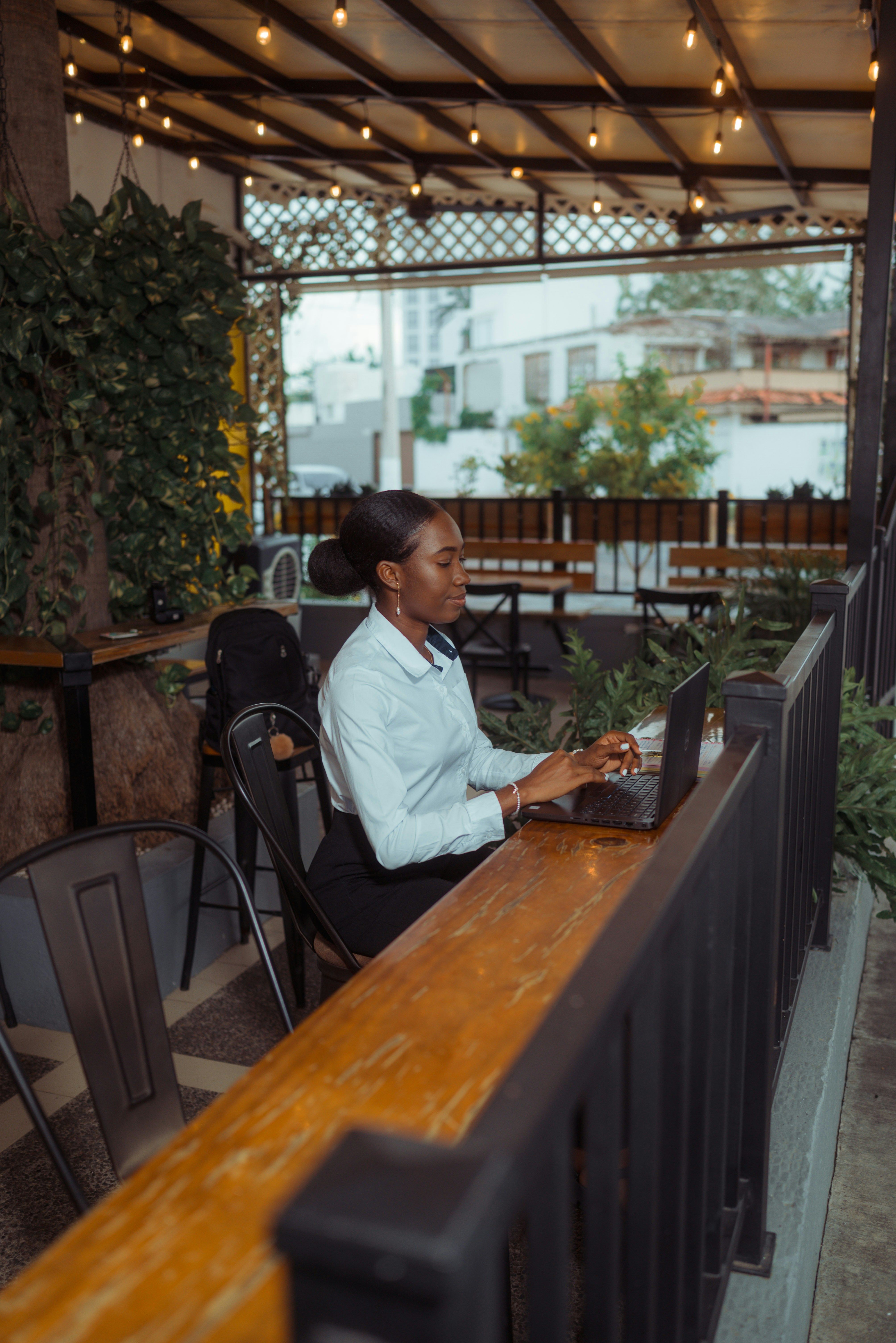 A woman in a white shirt works on a laptop at a wooden counter in an empty restaurant patio, with string lights and plants visible in the background.