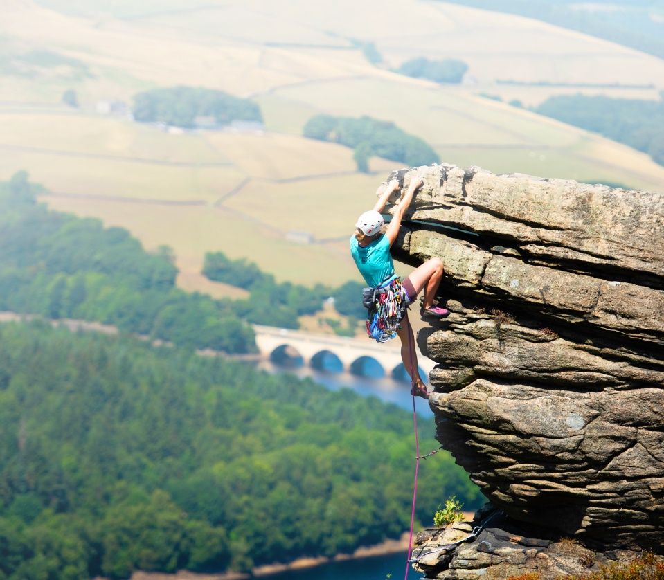 A climber in a helmet hanging from an overhanging gritstone edge, with a reservoir and viaduct in the valley below