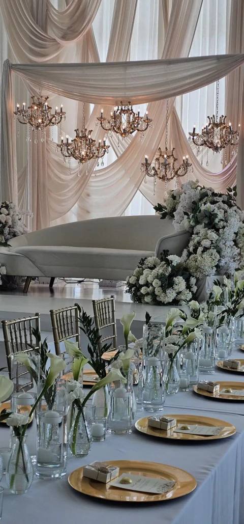 White curved lounge sofa framed by white hydrangeas and roses, beneath blush swag drapery and crystal chandeliers, behind a head table with calla lilies.