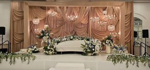 Stage with a sculpted champagne fan-pleat backdrop, crystal globe chandeliers and a canopy of white and blue florals above a curved white sofa.