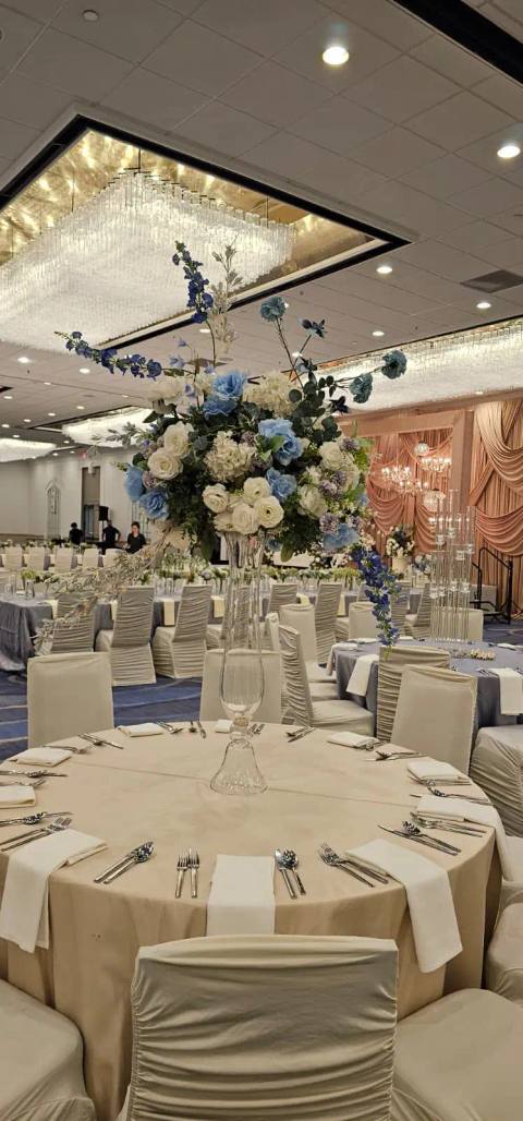 Tall centerpiece of blue and ivory roses and hydrangeas on a crystal stand, above a round table set in ivory linen in a ballroom under crystal chandeliers.